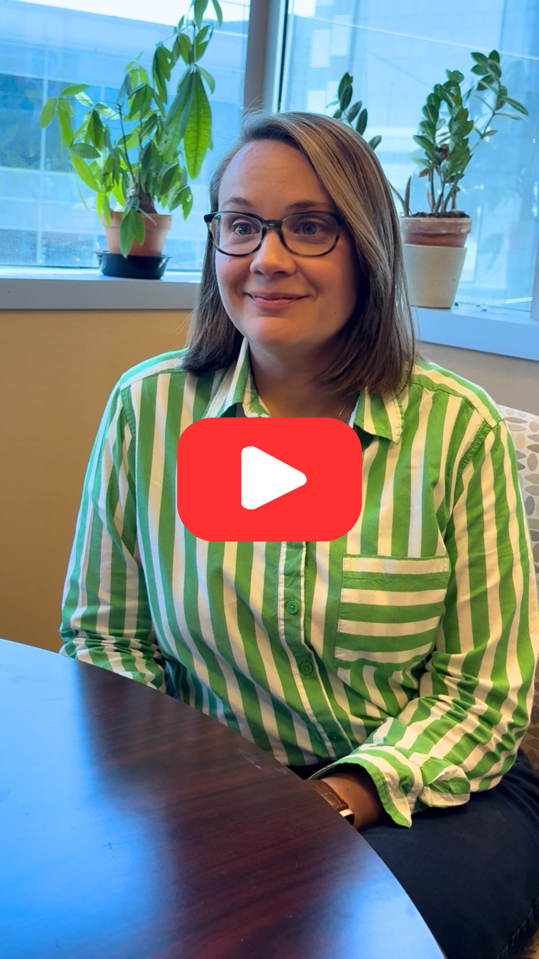 Ali Kowitz sits in her office speaking with someone off camera. A red YouTube Play button is at center of image.