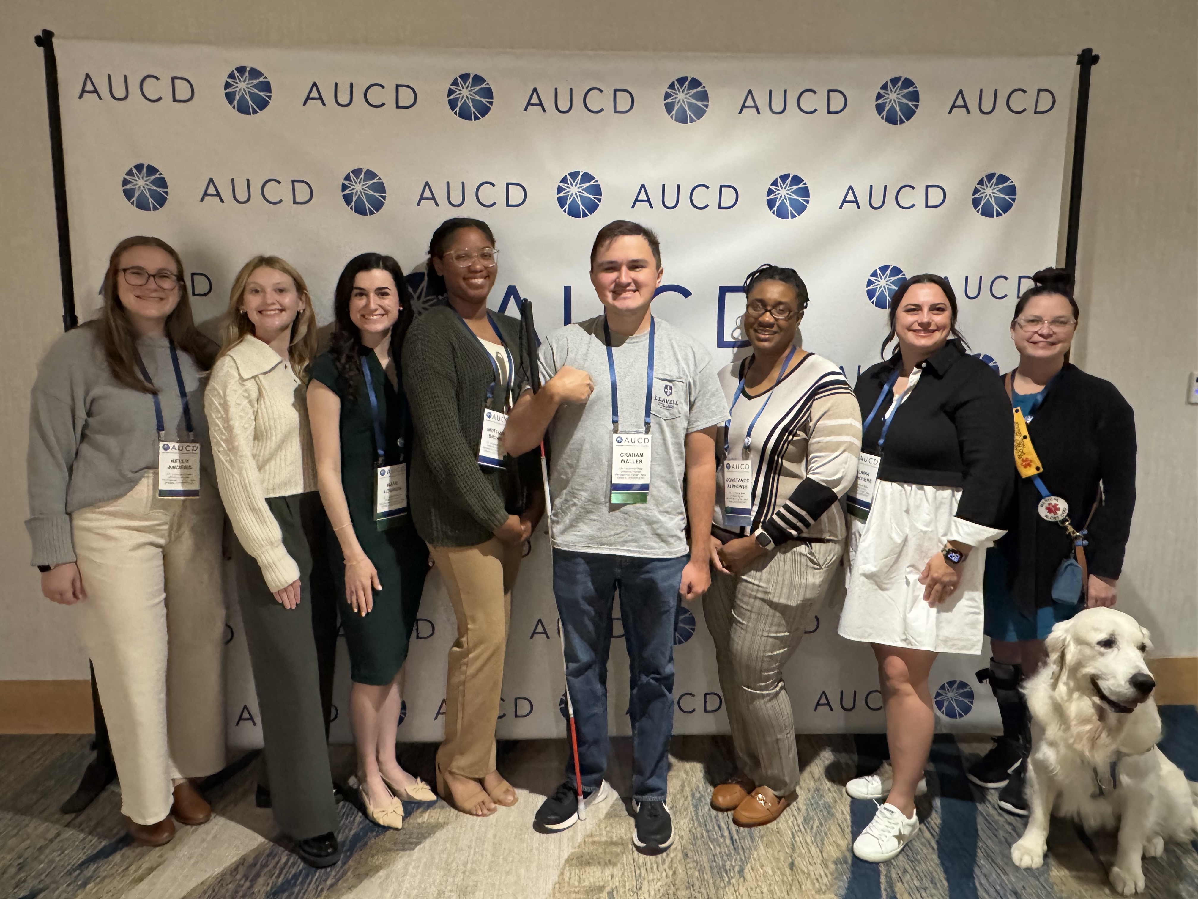 8 LEND scholars pose together in front of a AUCD banner. One scholar has a white cane and another has a service dog.