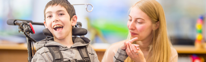 Woman helping small boy in motorized wheelchair inside a room that looks like a classroom