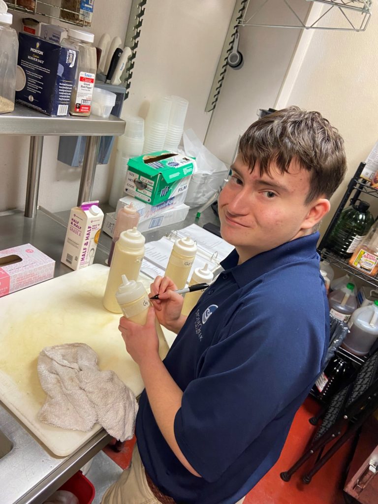 Teenage boy smiles as he prepares food in restaurant kitchen.