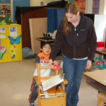 Woman guides young boy with deafblindness as he pushes his desk chair in the classroom.