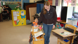 Woman guides young boy with deafblindness as he pushes his desk chair in the classroom.