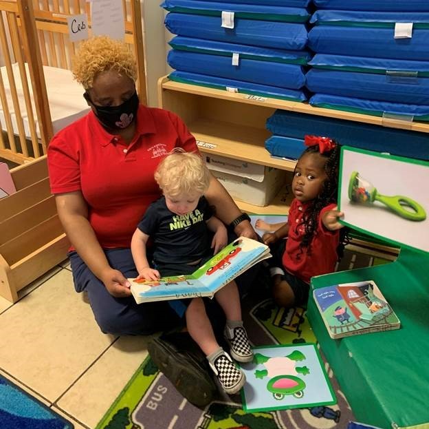 EHS-CCP teacher reading with toddler on her lap while another toddle holds book up to show the camera what she's reading.
