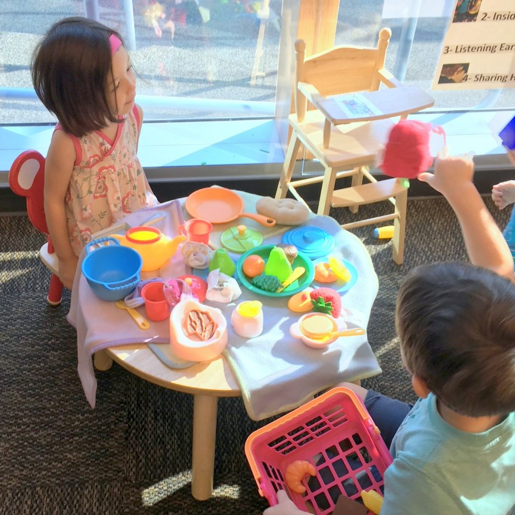 Two toddlers sit at a small table set with dozens of plastic food and cooking items while one boy waves a teapot in the air.