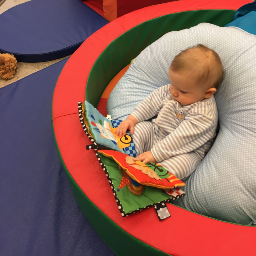 Infant about 4 months old reads picutre book while sitting up against plush support.