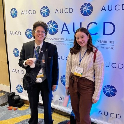 Margaret Shepard dressed professional and wearing an AUCD conference badge smiles next to Ivanova Smith dressed in a tie and holds a coffee cup. They stand in front of an AUCD banner.