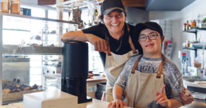 Young man with Down syndrome wearing an apron stands behind the counter of a cafe. Next to him stands an older man smiling also wearing an apron.