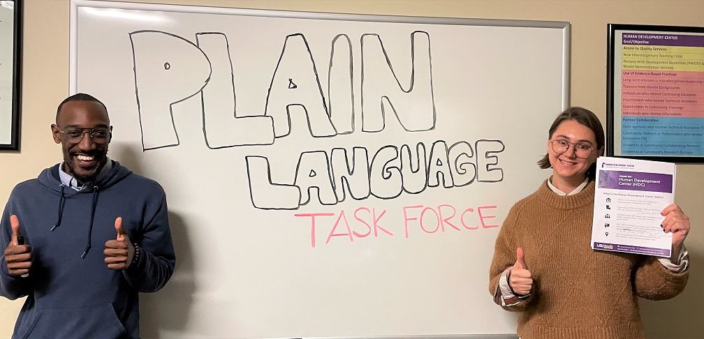 Will Johnson smiles with two thumbs up standing next to Jolie Robichaux holding her right thumb and document in plain language document in her left hand. Behind them is a white board with the text written in bubble letters: "Plain Language Task Force."