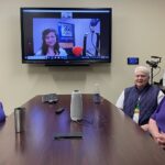 Rose Angelocci, Mary LeBlanc, and Loretta Brehm sit around a conference table in front of a screen that features the video feed of WWNO's Managing Producer Alana Schreiber during their interview. Rose wears a purple collard Krewe of King Arthur shirt, Mary wears a purple vest over a white t-shirt with the purple, green, and gold Mardi Gras colors, and Loretta wears a Krewe of King Arthur t-shirt with the logo in white. The Krewe of King Arthur logo is King Arthur wearing a crown with his hand on the holt oh his sword. Behind him are trees and a castle.