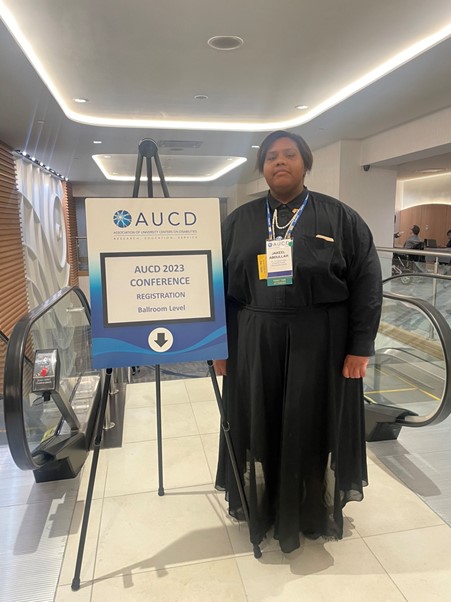 Jakeel wearing conference name-badge, a black blouse, and black floor-length skirt stands next to the AUCD 2023 Conference sign.