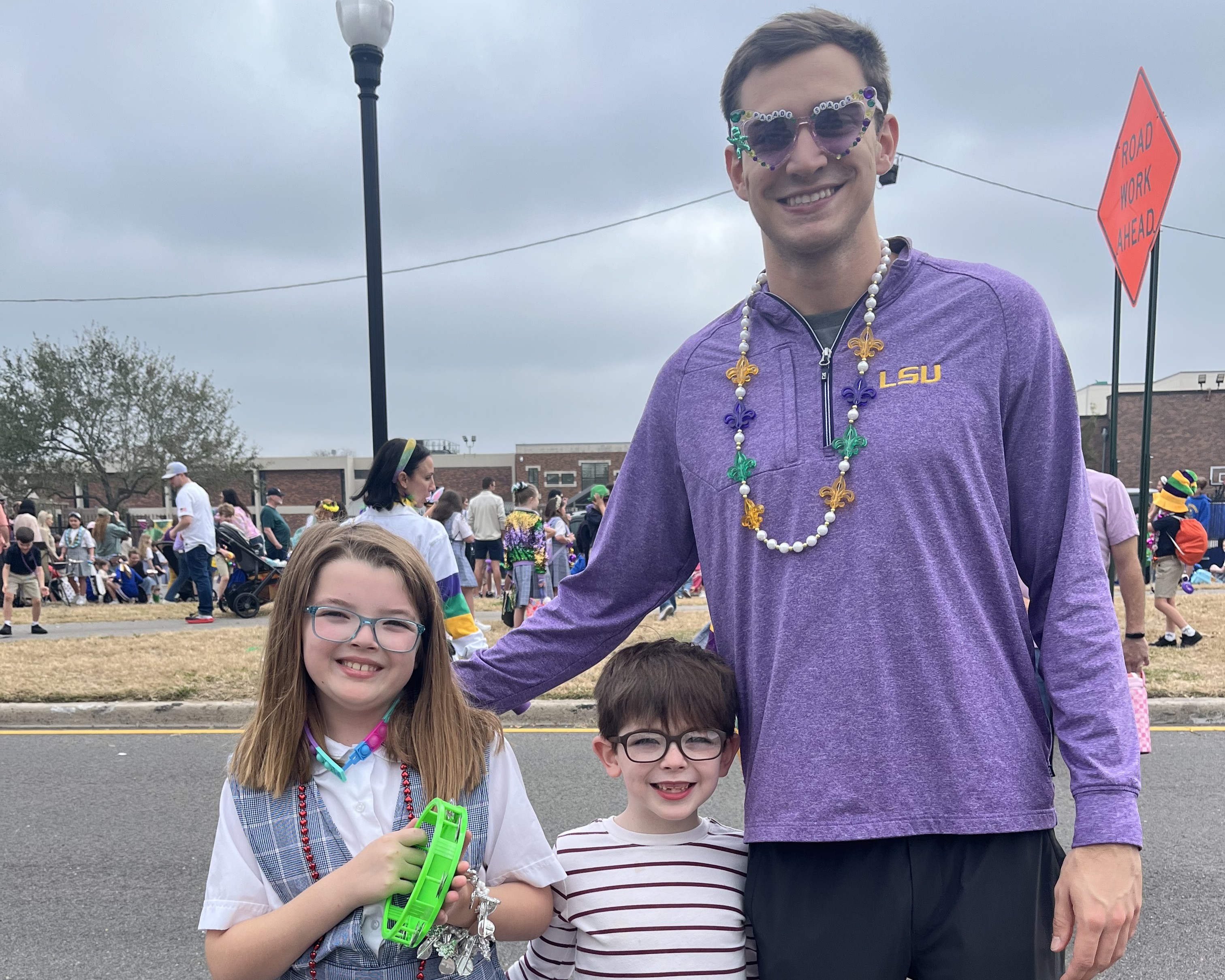 Scholar wearing purple LSU shirt adn mardi gras beads stands smiling with two young children at their school parade. 